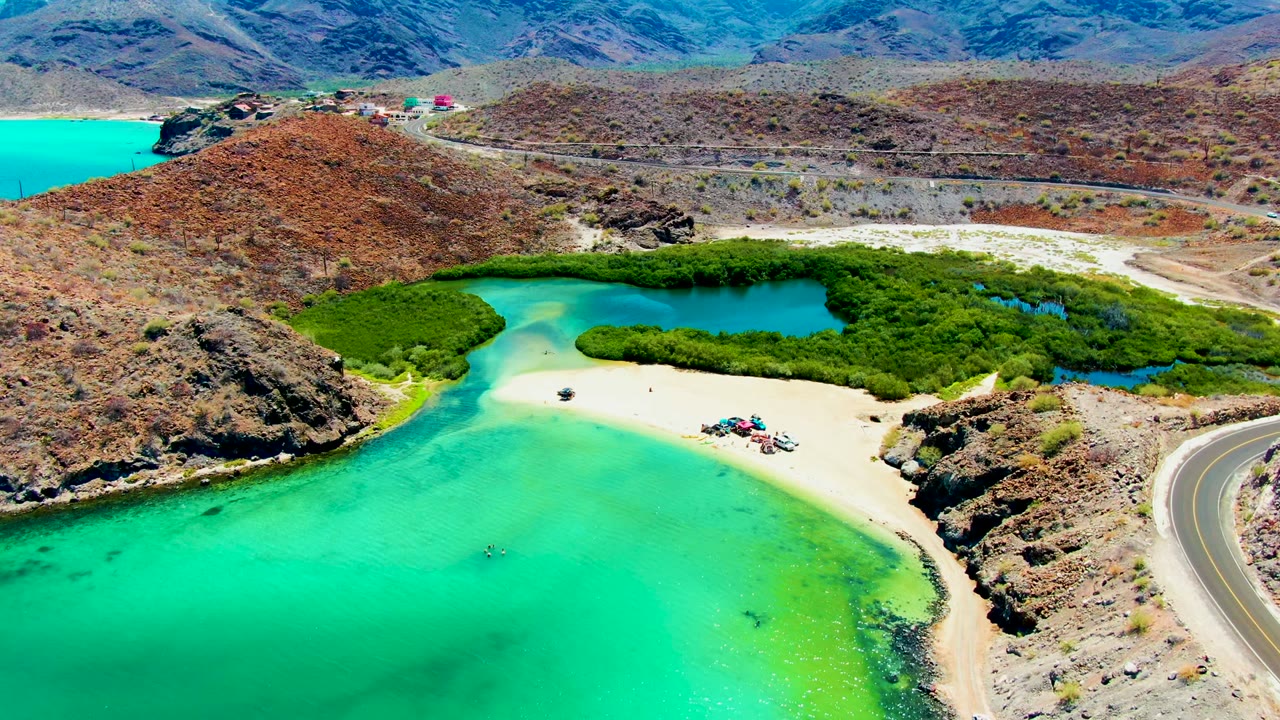 Aerial frame over Playa Santispac showing turquoise water in front, a pale sand beach with tiny people and umbrellas, a curving road at right, green mangroves, and dry hills beyond.