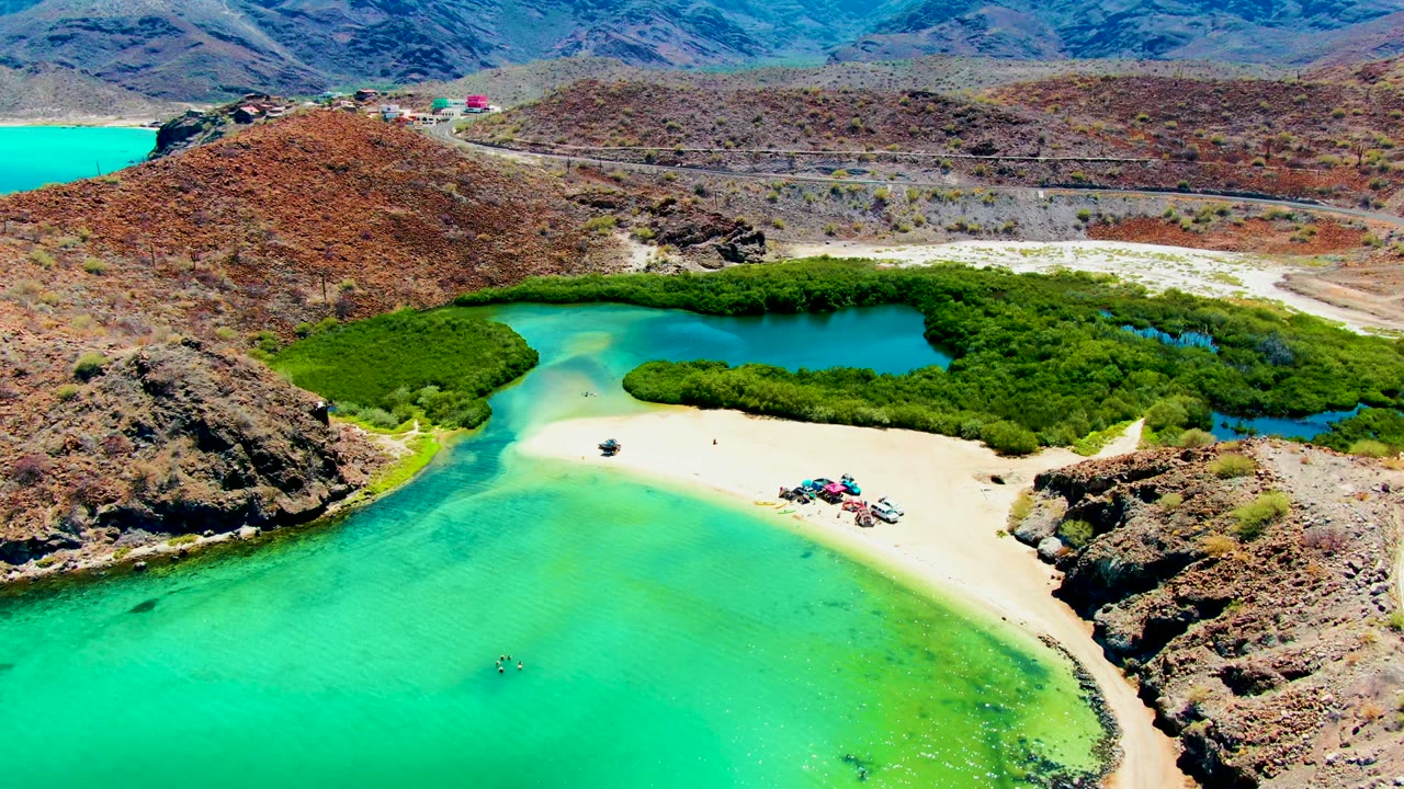Playa Santispac aerial frame with bright turquoise water across the foreground, beach umbrellas and small figures on the sand, mangroves wrapping a lagoon, and a road partly visible at the far right.