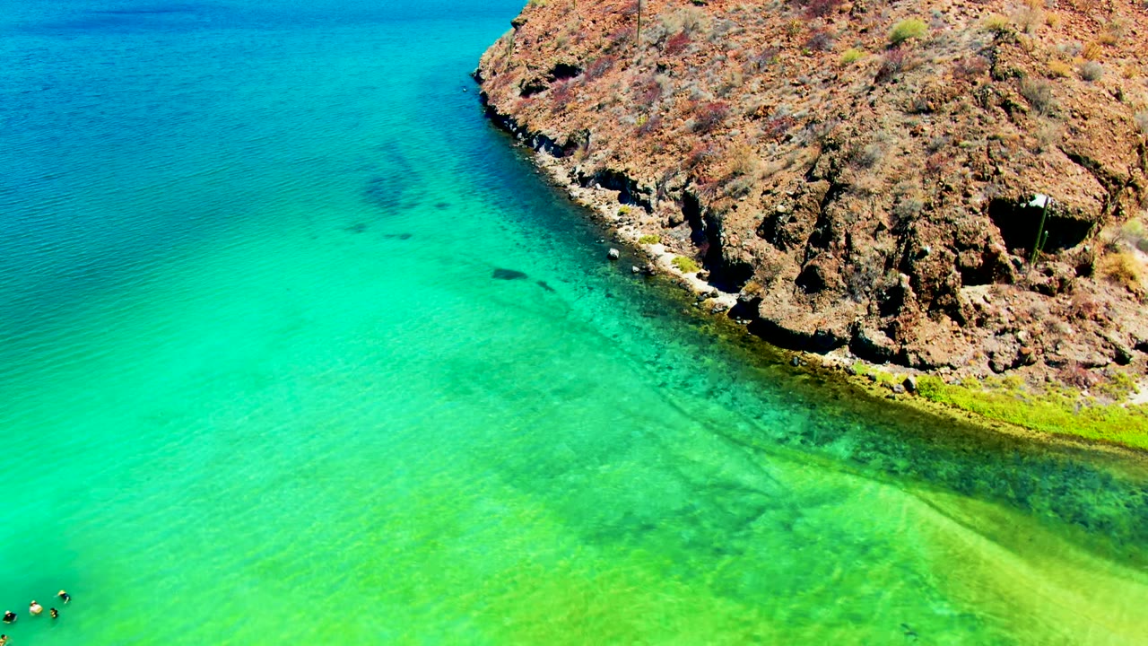 Aerial view of Playa Santispac with a rocky brown shoreline on the right, bright turquoise water on the left, and tiny swimmers partly visible at the lower left edge.