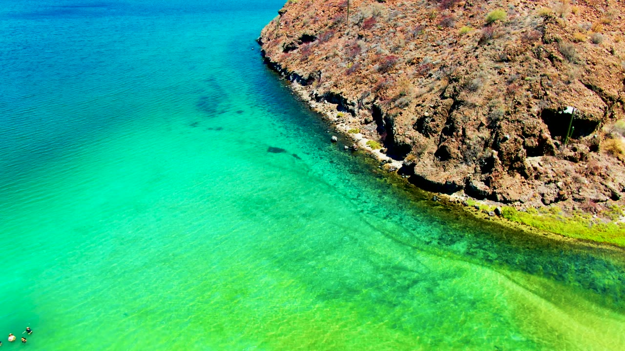 Turquoise coastal water and rocky headland with tiny swimmers near the lower left.