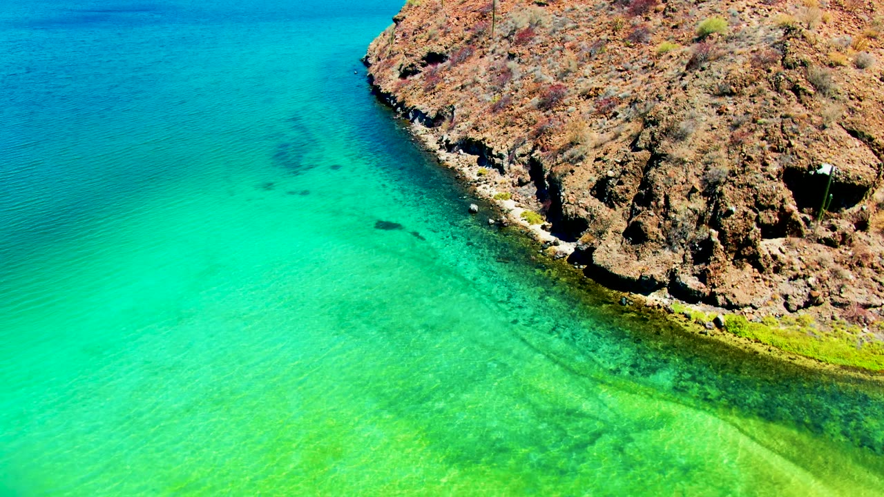 Aerial view of Playa Santispac where the rocky shore curves across the upper right and clear turquoise water shifts to bright green near the shallows, with no visible people.