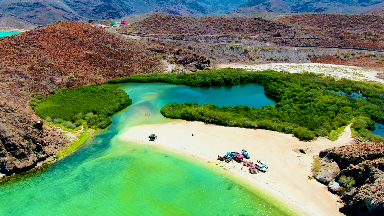 Aerial frame from Playa Santispac, Baja California Sur, showing turquoise water at lower left, pale sand with vehicles and tiny people near the center, dense green shoreline vegetation, and brown hills beyond a road.