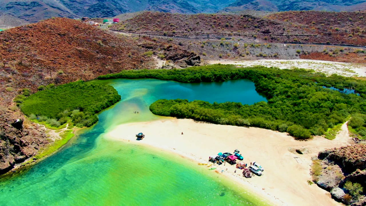 Aerial beach view with vehicles on pale sand beside green vegetation and clear blue-green water.
