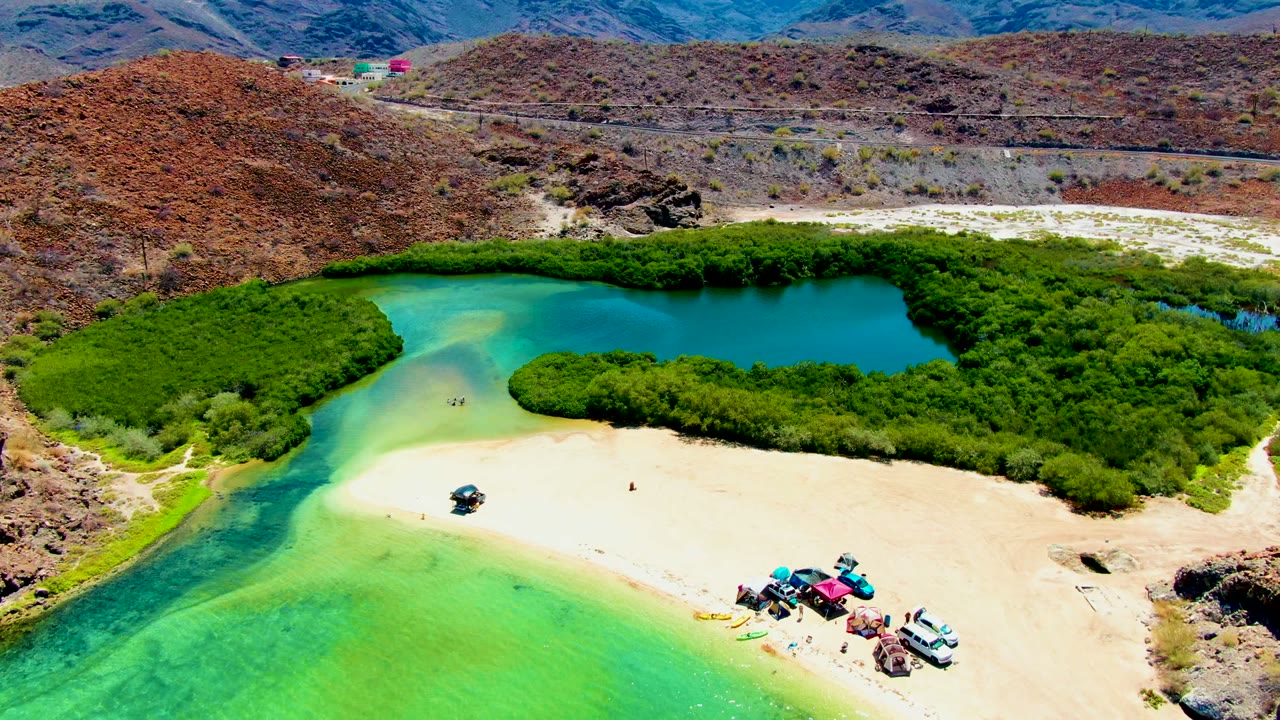 At 0:02, the aerial view places the Playa Santispac shoreline across the frame, with bright turquoise water in the foreground, vehicles and beach items on the sand, green vegetation around the lagoon, and a road below the hills.