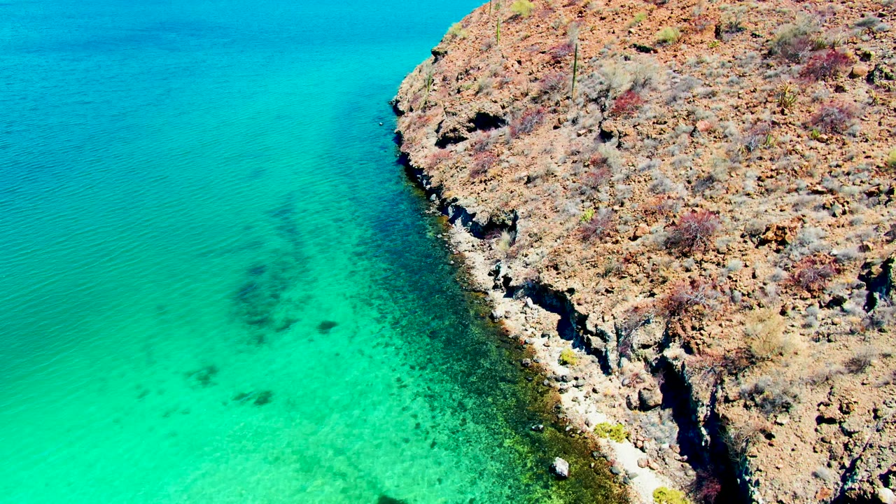 Aerial view of Playa Santispac turquoise water meeting a rugged dry rocky shore with sparse vegetation and no visible people.