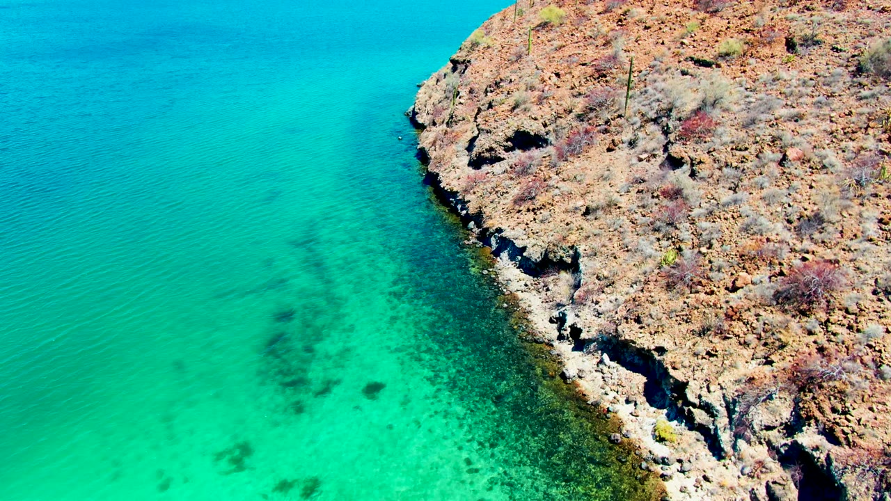 Aerial view of Playa Santispac with a rocky point entering from the right and turquoise coastal water spreading across the left, with no visible people.