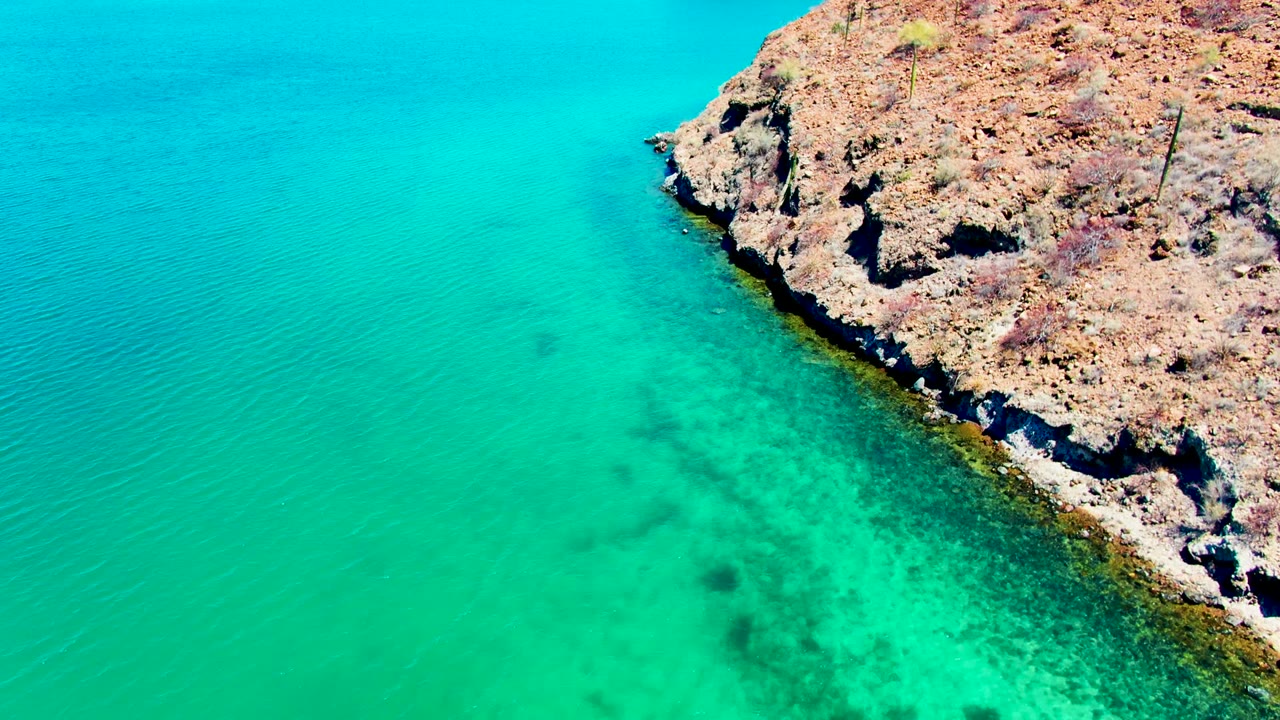 Aerial view at Playa Santispac, Baja California Sur, showing turquoise water beside a tan rocky shoreline with sparse dry vegetation and no visible people.
