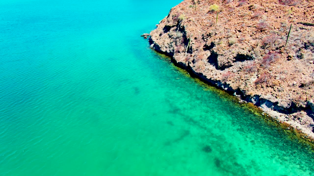 Aerial view at Playa Santispac where a rocky point enters from the upper right into bright turquoise water with faint ripple lines and no visible people.