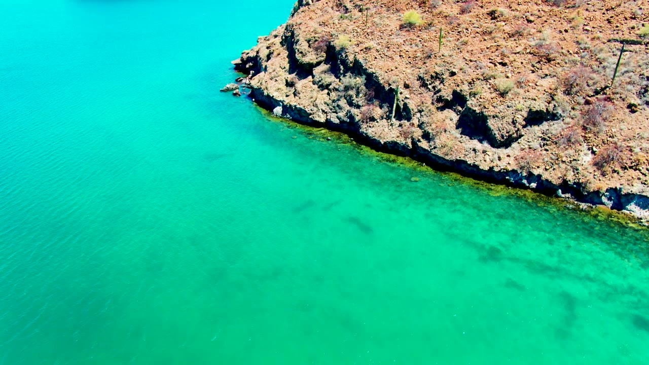 Aerial view of Playa Santispac rocky shoreline above clear turquoise water, with sparse scrub on the dry land and no visible people.