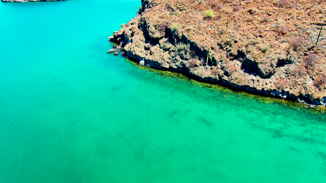 Aerial view over Playa Santispac water with a rocky point across the top of frame and darker submerged patches visible below the surface, with no visible people.