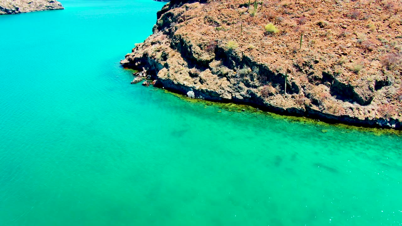 Aerial view of turquoise water meeting a rugged rocky point at Playa Santispac, Baja California Sur, with no visible people.