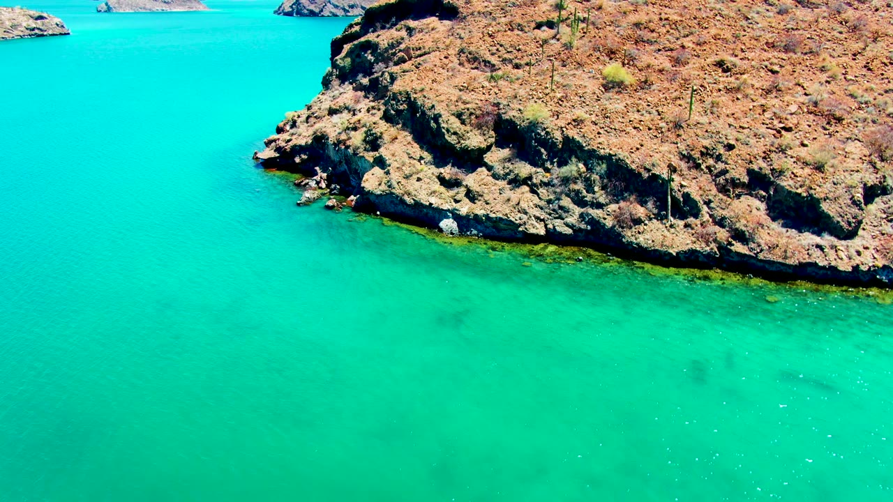 Aerial view at Playa Santispac with turquoise sea filling most of the frame, a rocky bluff along the upper edge, and no visible people.