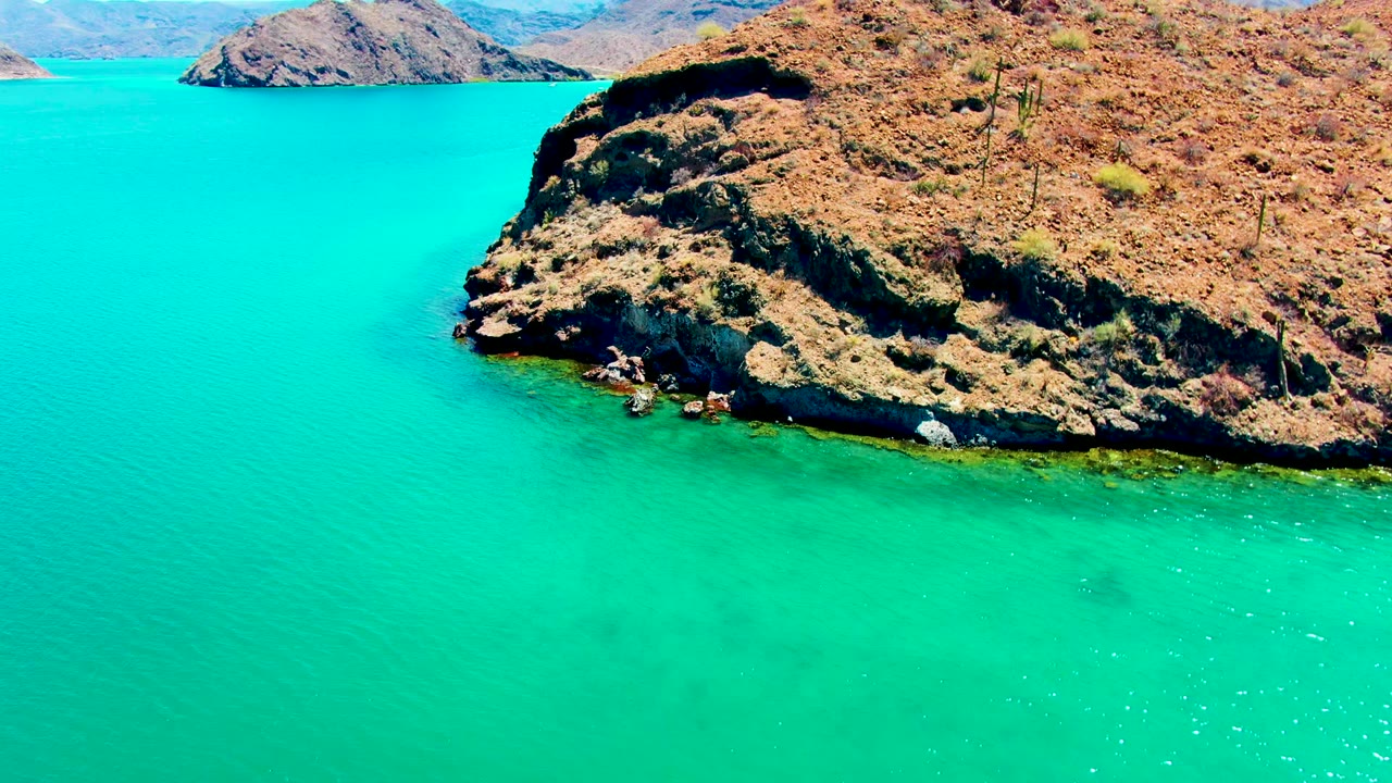 Opening aerial frame over Playa Santispac shows clear turquoise water, a dry rocky headland with sparse cactus, distant hills, and no visible people.