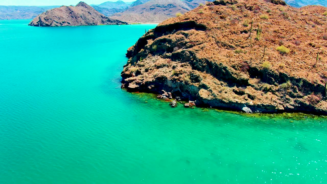 At two seconds, the dry cliff edge and cactus-dotted slope sit above bright turquoise sea, with layered mountain ridges in the background and no visible people.
