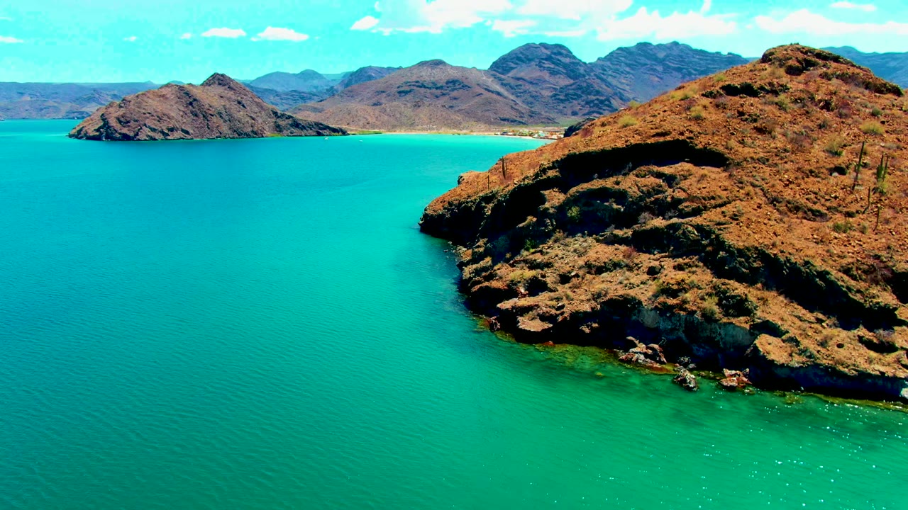 Opening aerial frame over Playa Santispac, Baja California Sur, with turquoise water around a brown rocky point, distant arid mountains, scattered clouds, and no visible people.