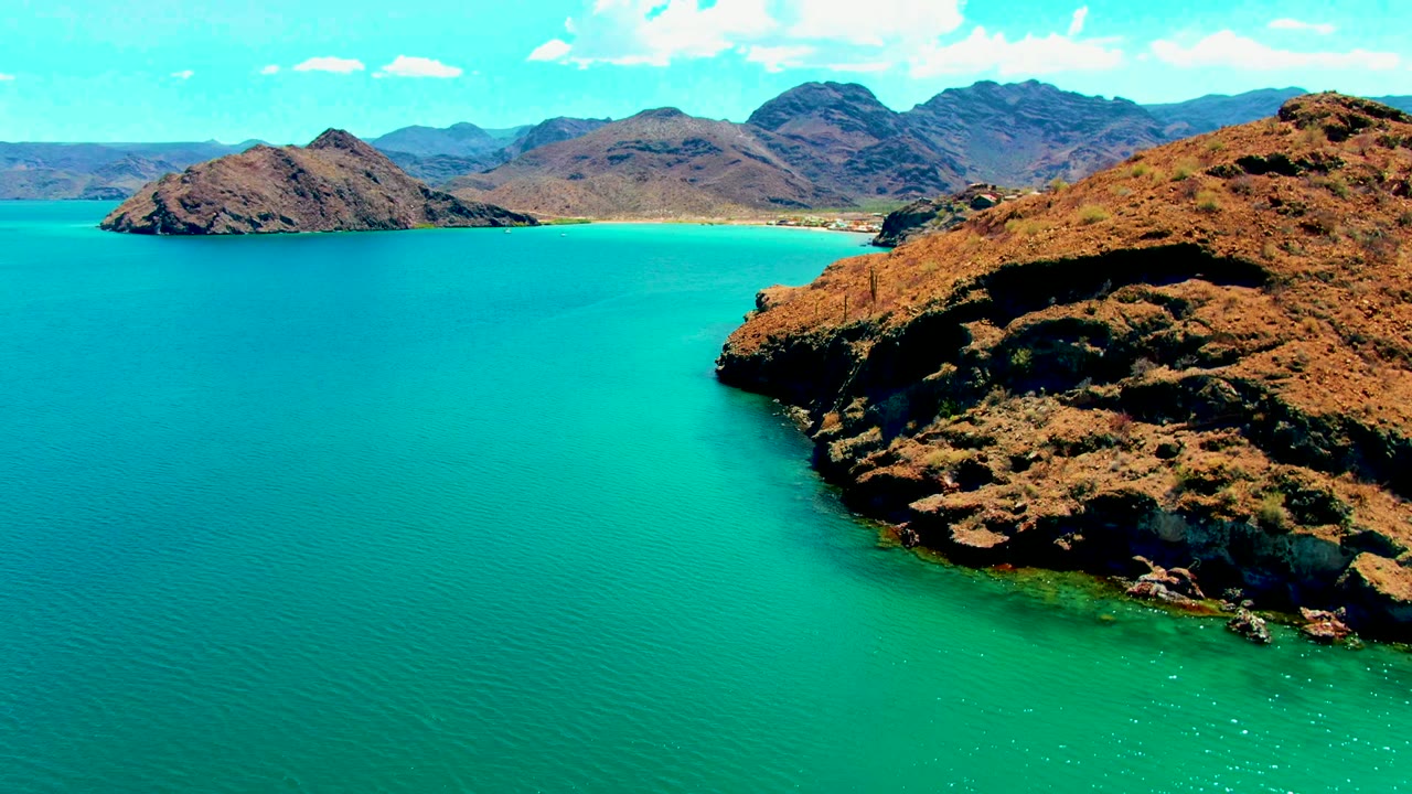 Aerial frame over Playa Santispac showing turquoise bay water, the rocky point cropped along the right edge, distant brown mountains, small white clouds, and no visible people.