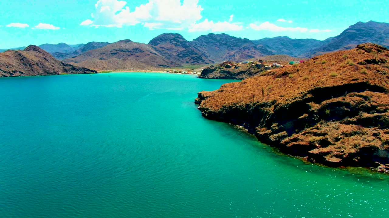 Opening frame of Playa Santispac shows turquoise water in the foreground, a dry rocky headland on the right, distant mountains, puffy clouds, and no visible people.
