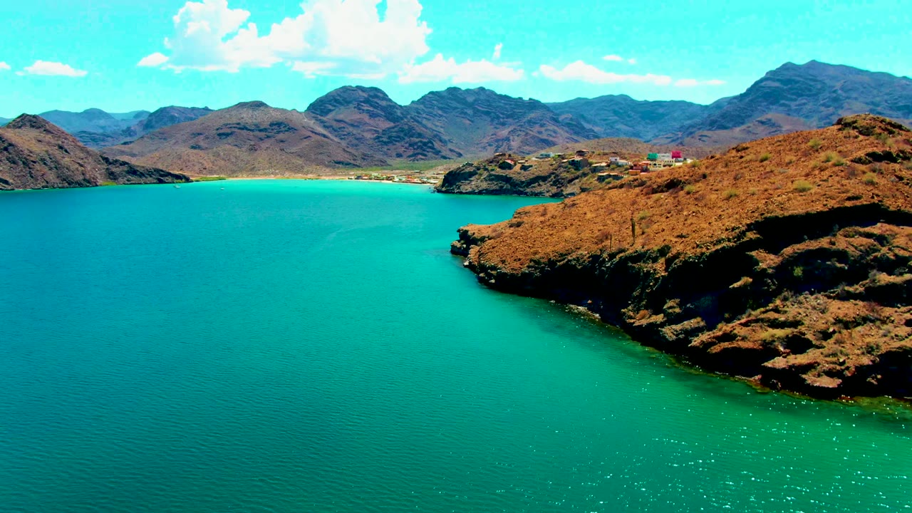 At 0:02, Playa Santispac appears as a wide turquoise bay with the rocky headland forming a diagonal right-side edge, mountain ridges in back, and no visible people.
