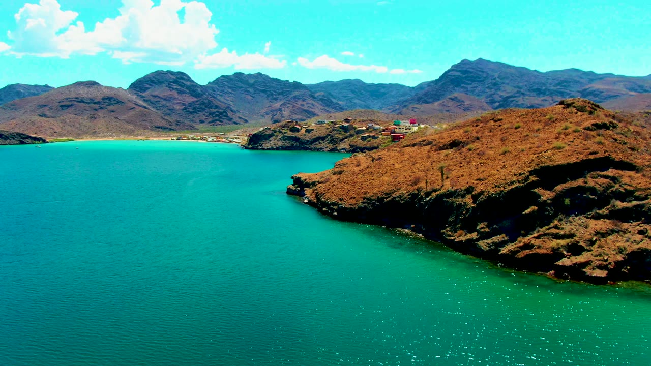 Opening aerial view of Playa Santispac with turquoise water in the foreground, a rust-brown rocky point at right, distant mountains under scattered clouds, and no visible people.