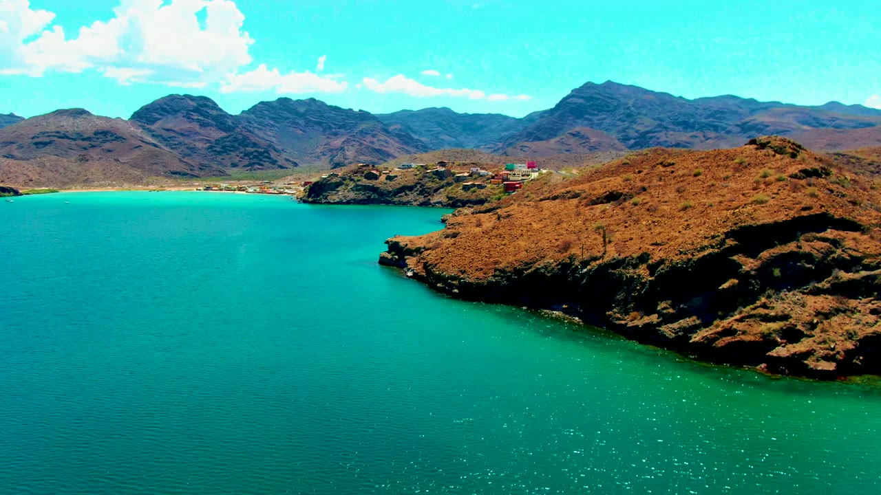 Playa Santispac aerial view with the rocky point pushed farther right, bright turquoise water across the lower frame, dry mountains along the horizon, and no visible people.