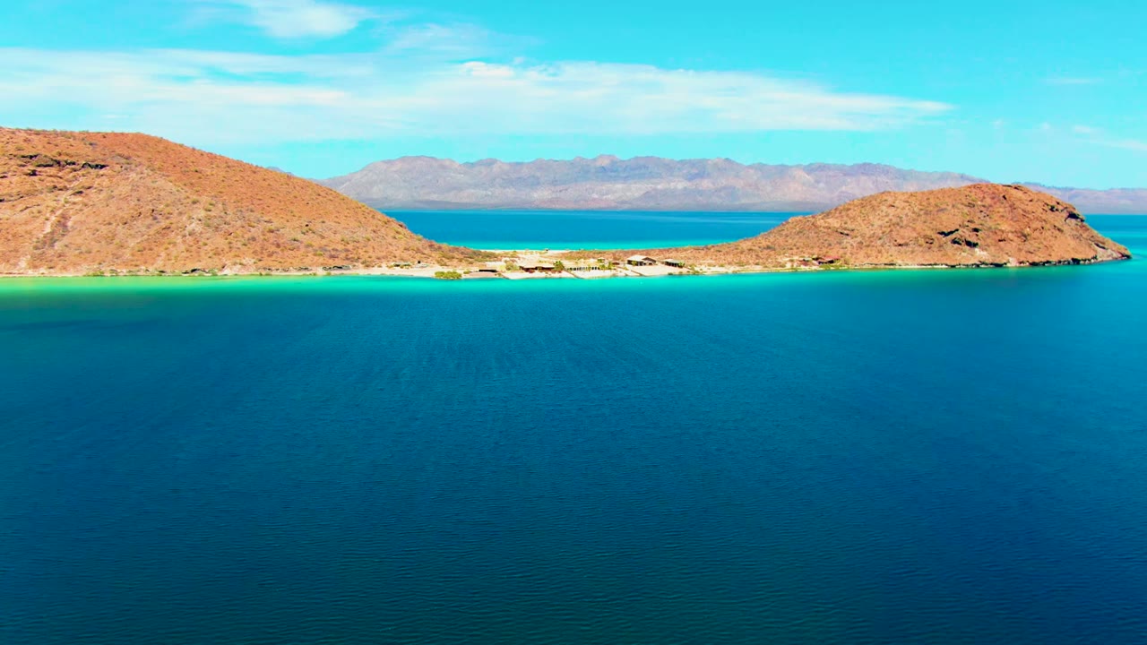At 0:00, Playa Santispac water fills the foreground below a narrow turquoise channel between rocky brown islands, with distant mountains and no visible people.