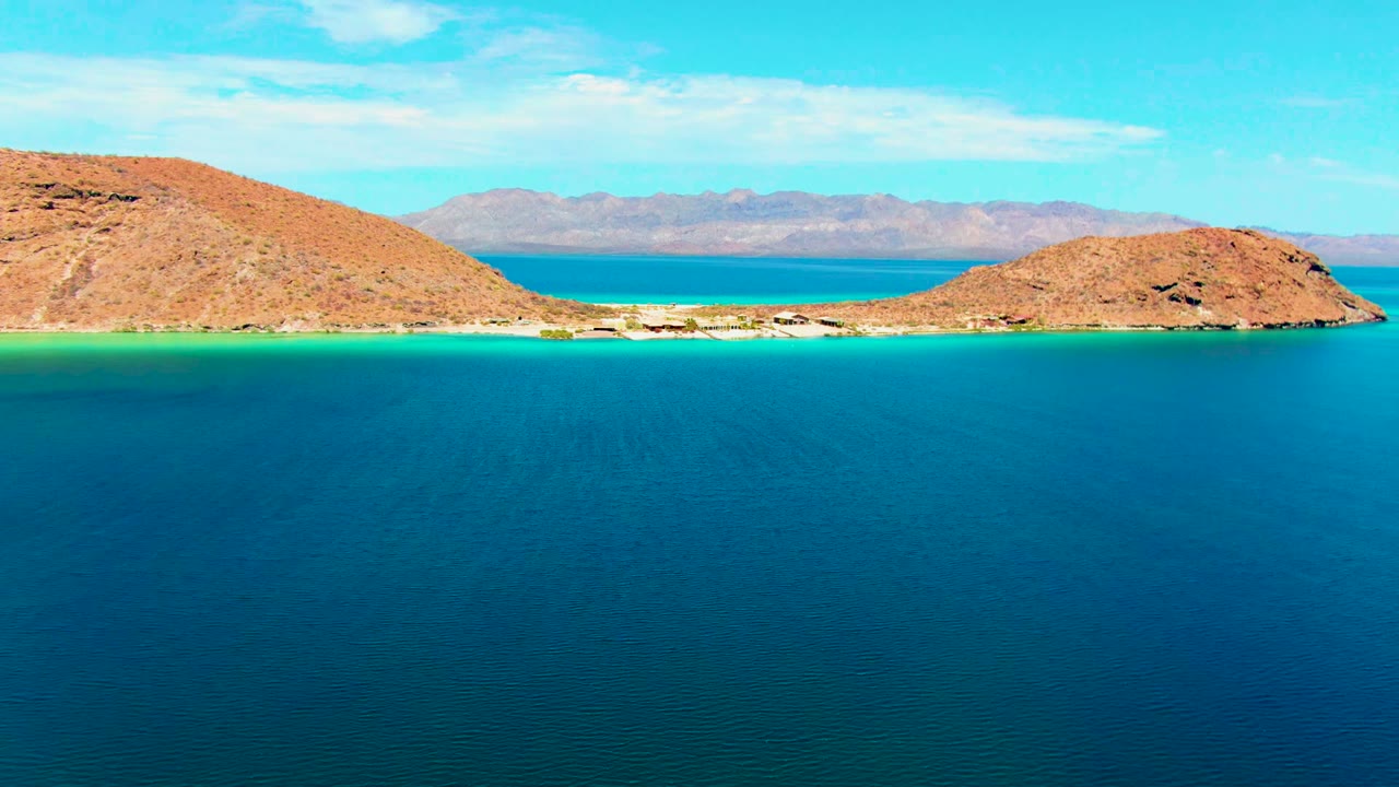 At 0:02, deep foreground water leads toward the narrow Playa Santispac channel, with rocky island hills on both sides and a pale mountain ridge beyond, no visible people.