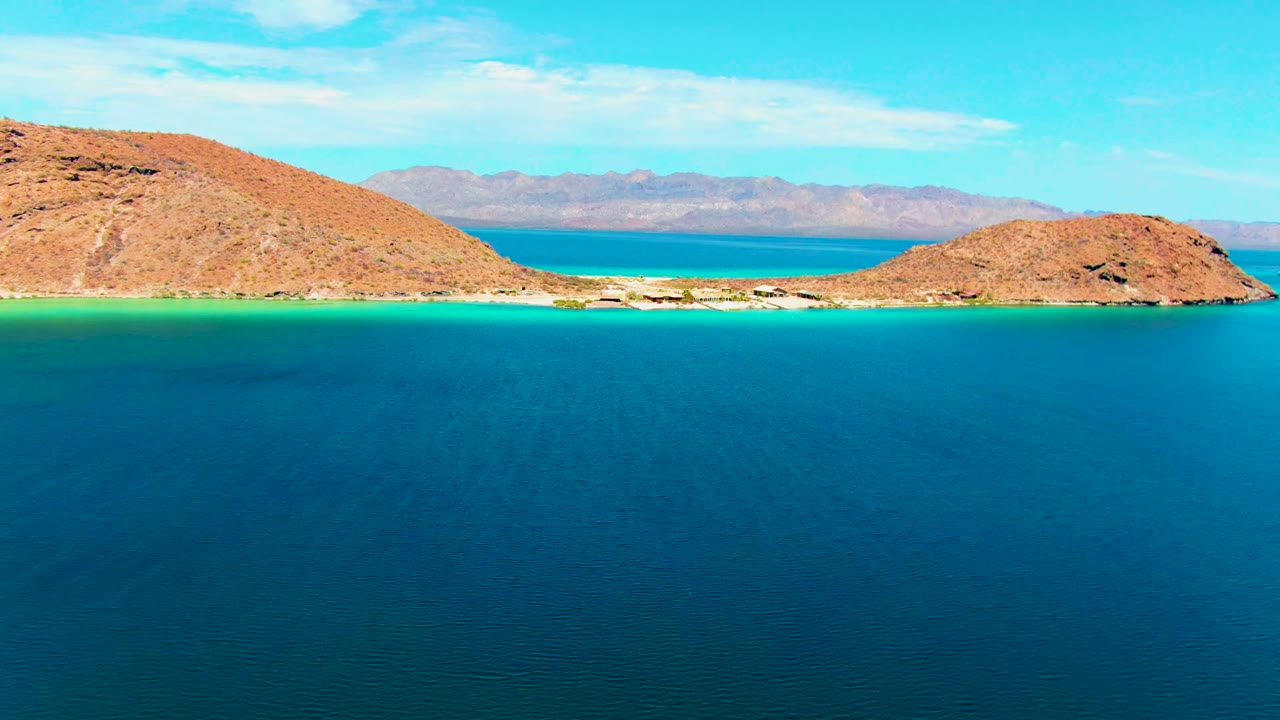 At Playa Santispac in Baja California Sur, deep blue water fills the foreground below rocky tan shores, turquoise shallows, distant mountains, and no visible people.