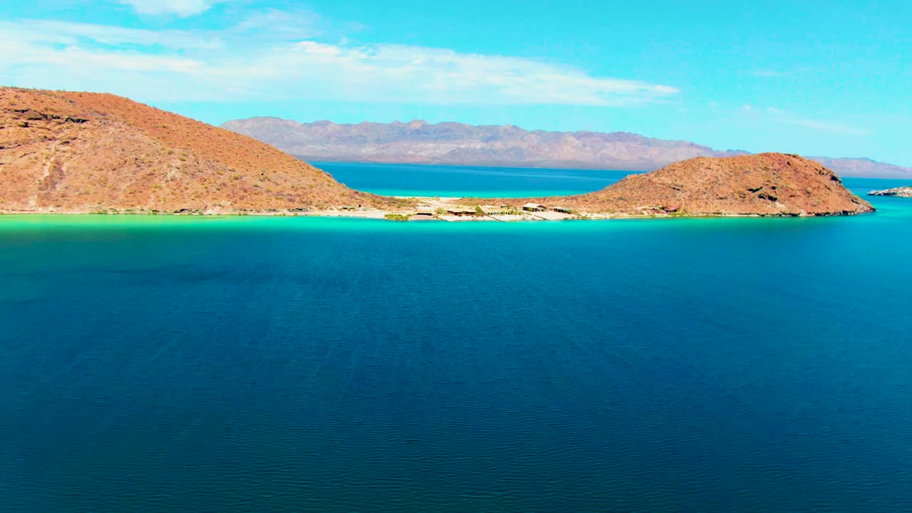 At 0:02, the aerial view keeps the Playa Santispac rocky shores centered around the narrow channel, with textured blue water in front and no visible people.