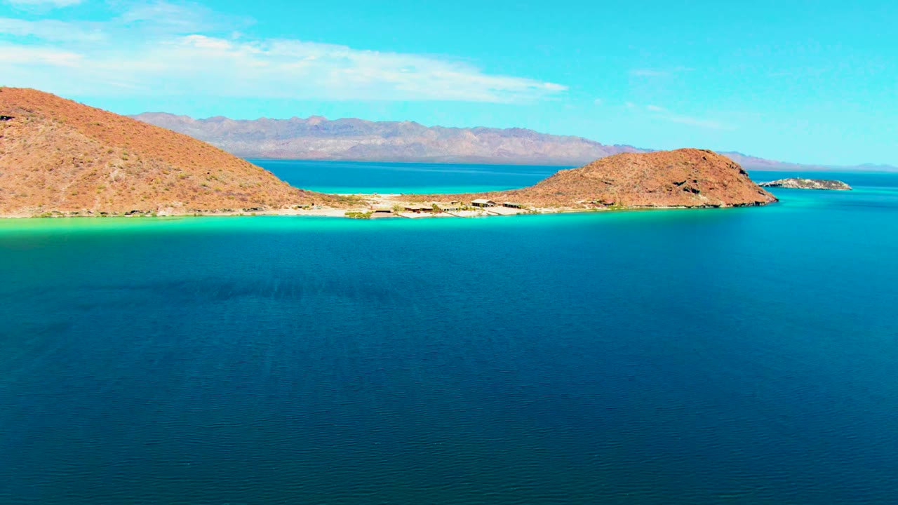 Wide aerial view at Playa Santispac showing deep blue water, turquoise shallows, a sandy strip between rocky hills, distant mountains, scattered clouds, and no visible people.