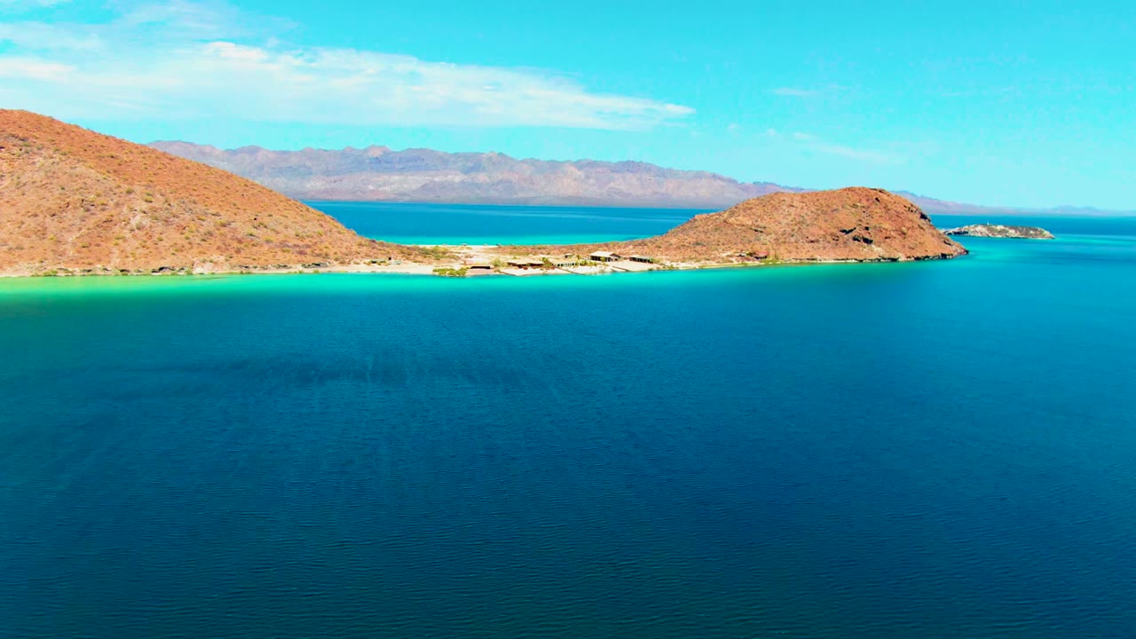 Aerial frame at Playa Santispac with blue foreground water, bright turquoise shoreline, two dry rocky hills enclosing the beach, distant mountain ridge, and no visible people.