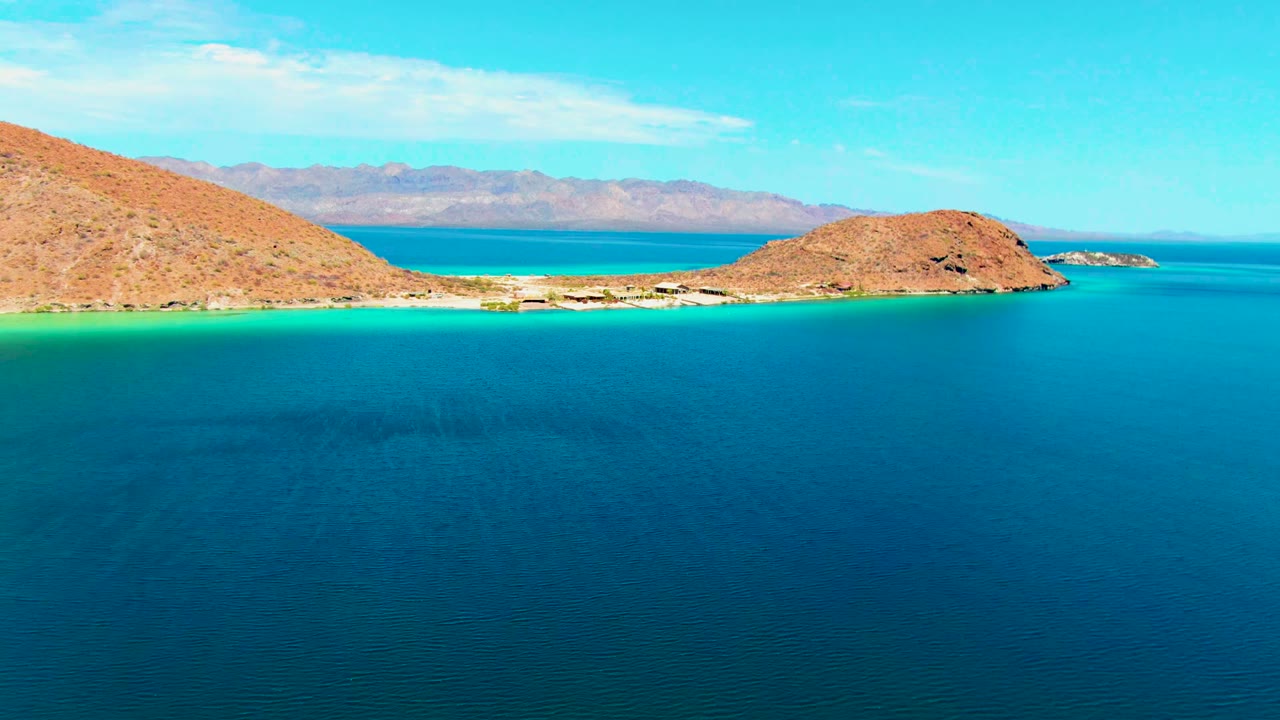 Playa Santispac aerial view with calm dark blue water in front, a pale sandy shore edged by turquoise shallows, dry hills, distant land across the bay, and no visible people.