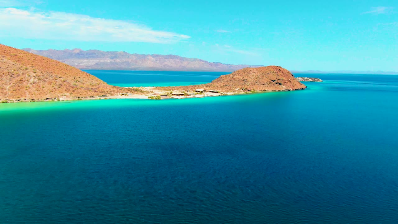Opening frame over Playa Santispac with deep blue foreground water, a turquoise shallows band at the beach, tan rocky hills, distant mountains, and no visible people.