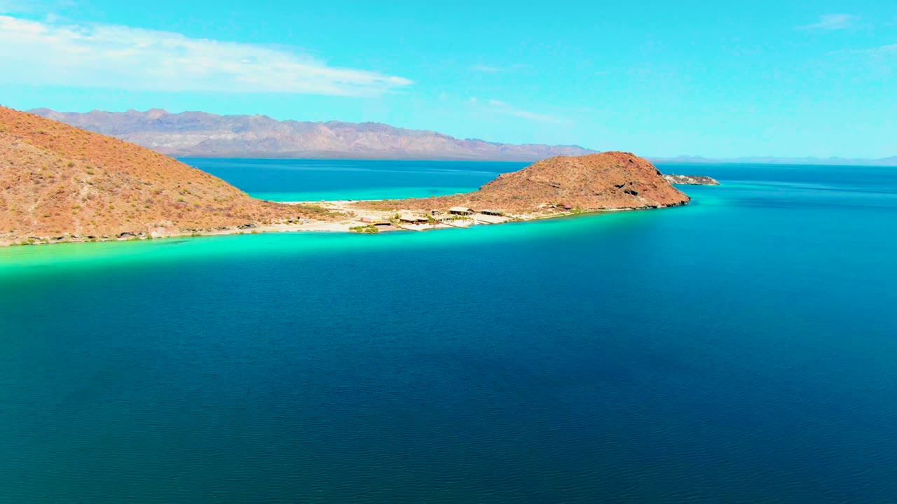 At 0:02, the rocky headland on the right and sloping hill on the left frame the Playa Santispac beach, with calm blue water in front and no visible people.