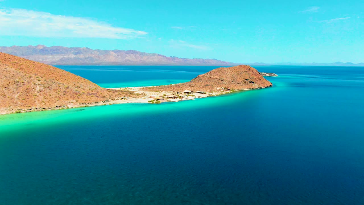 Wide aerial view of Playa Santispac with arid hills at left, sandy shoreline below, turquoise shallows, deep blue bay water, distant mountains, and no visible people.