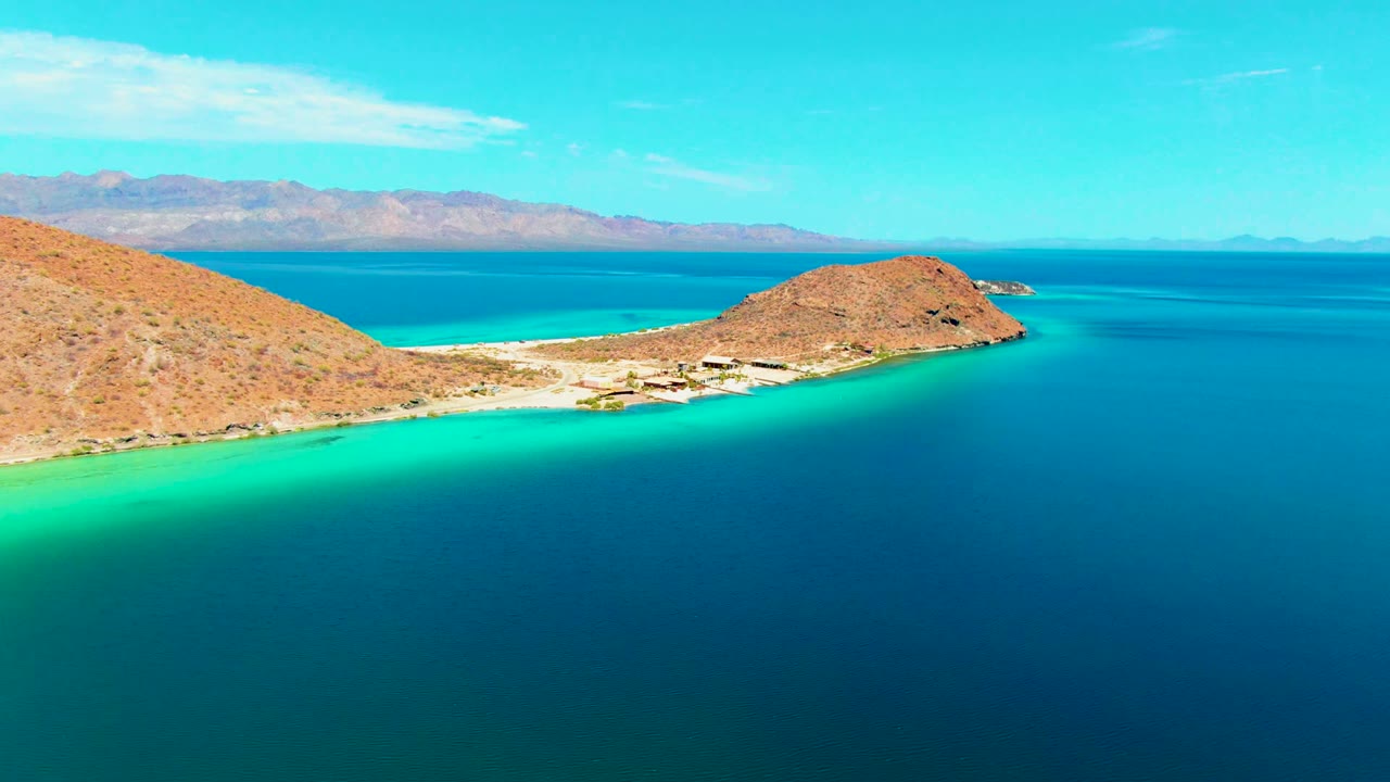 Playa Santispac aerial view with the left hillside, small beach structures near the sand, bright turquoise water near shore, darker bay water beyond, and no visible people.