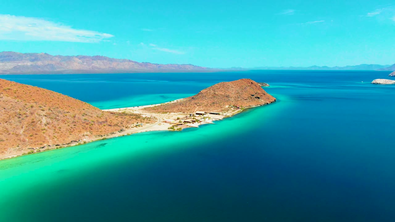 Opening aerial frame over Playa Santispac showing an arid peninsula, pale beach, turquoise shallows, deep blue bay, distant mountains, bright sky, and no visible people.