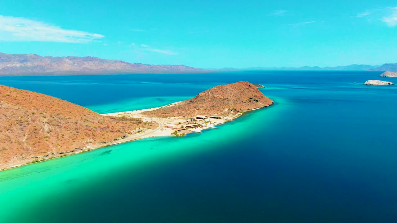 Wide aerial frame over Playa Santispac showing the rounded peninsula point, pale shoreline, dark blue open water, distant mountain ridge, sparse clouds, and no visible people.