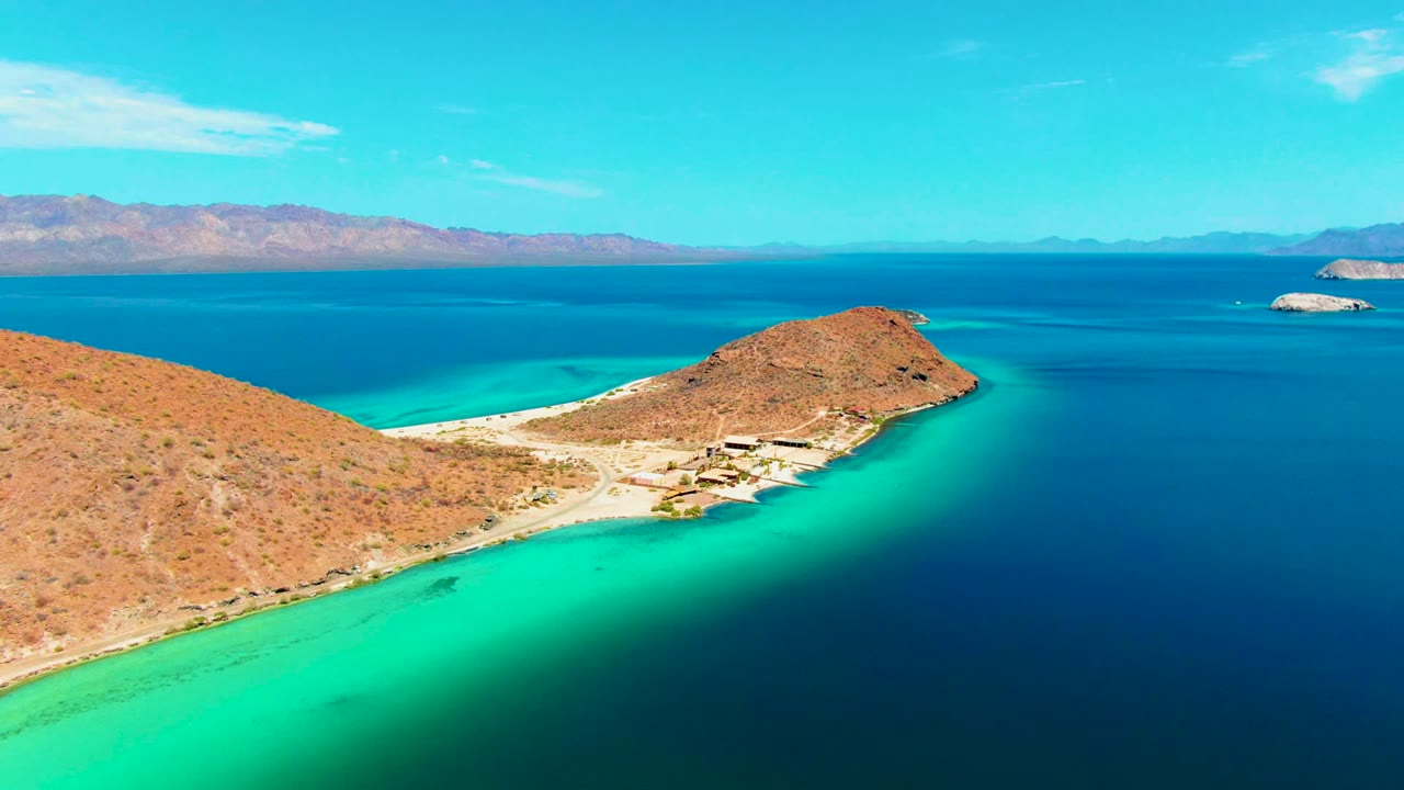 Aerial view over Playa Santispac with a rocky tan slope, bright turquoise shallows, deep blue ocean, distant mountains, and no visible people.