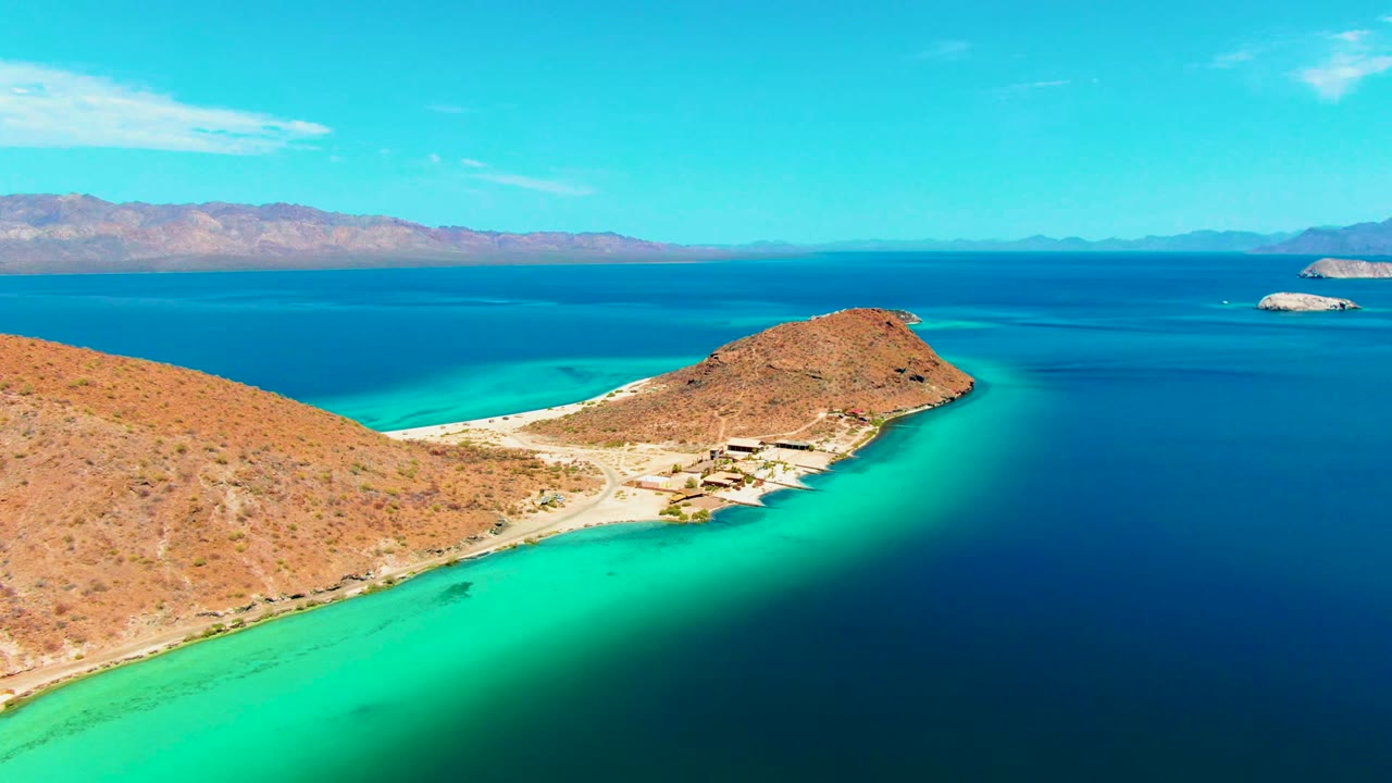 Playa Santispac coastline with the desert headland more centered, pale sand edging the cove, a small cluster of beach structures, blue water, and no visible people.