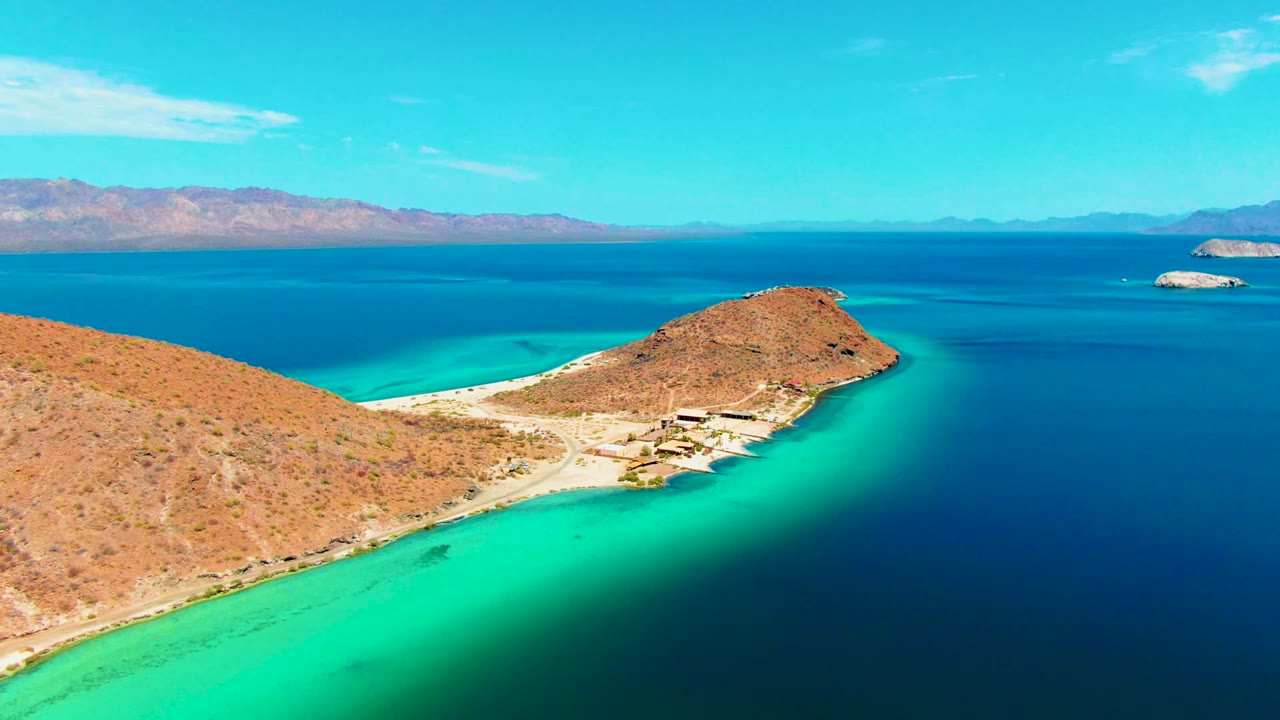 Wide Playa Santispac aerial with tan rocky land crossing the lower frame, turquoise shallows along the beach, dark blue open water, distant mountain ridges, and no visible people.