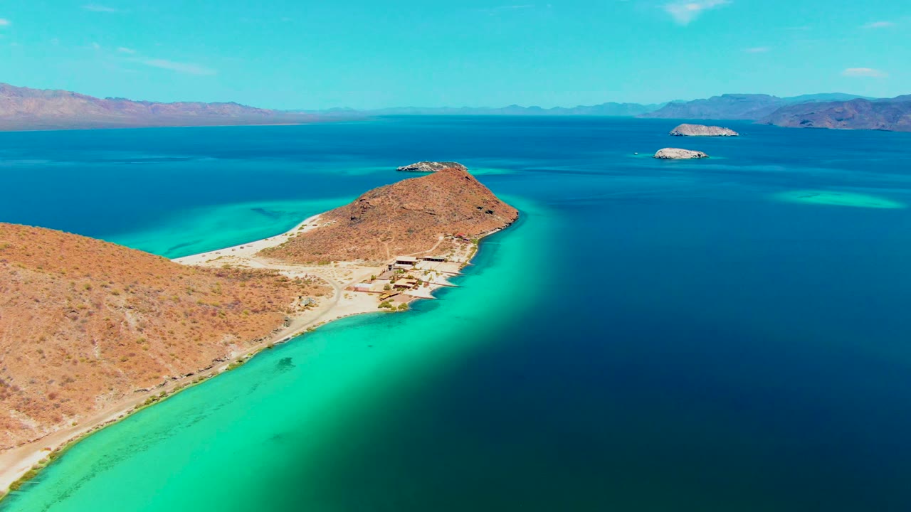 Aerial frame over Playa Santispac showing a rocky desert peninsula, a pale shoreline road, turquoise shallows, deep blue water, distant mountains, and no visible people.