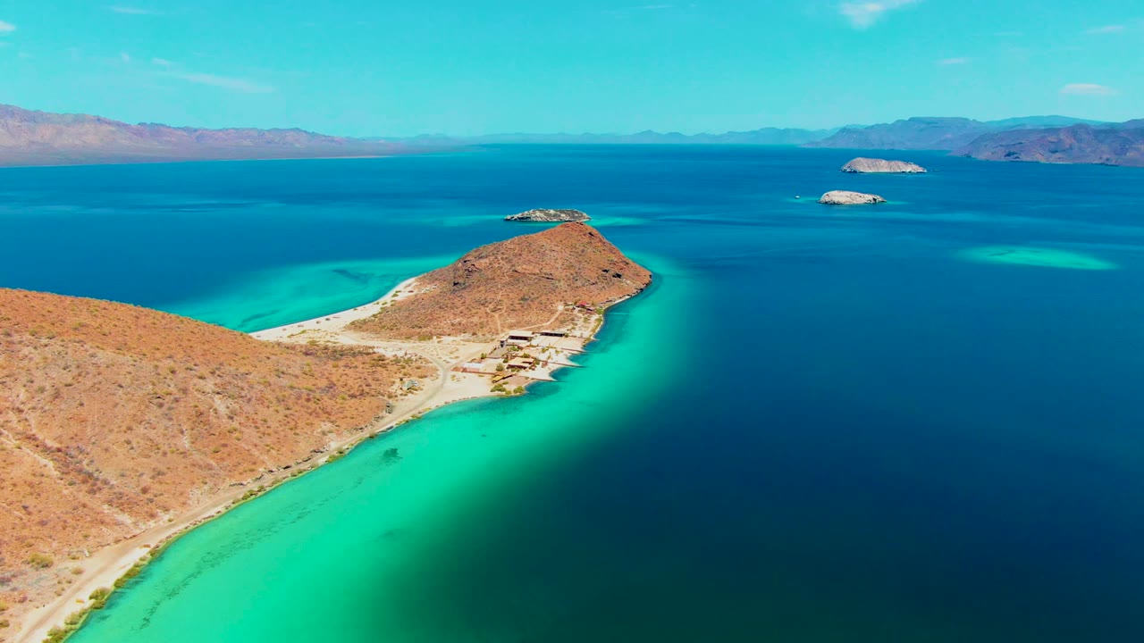 Aerial view over Playa Santispac with an arid peninsula, turquoise shallows, deeper blue water, small distant islands, mountains on the horizon, and no visible people.