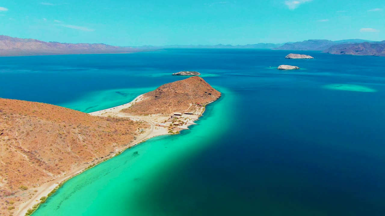 Playa Santispac aerial still with the sandy point and low desert hill centered near the frame, bright shallow water on the left, dark open water on the right, islands beyond, and no visible people.