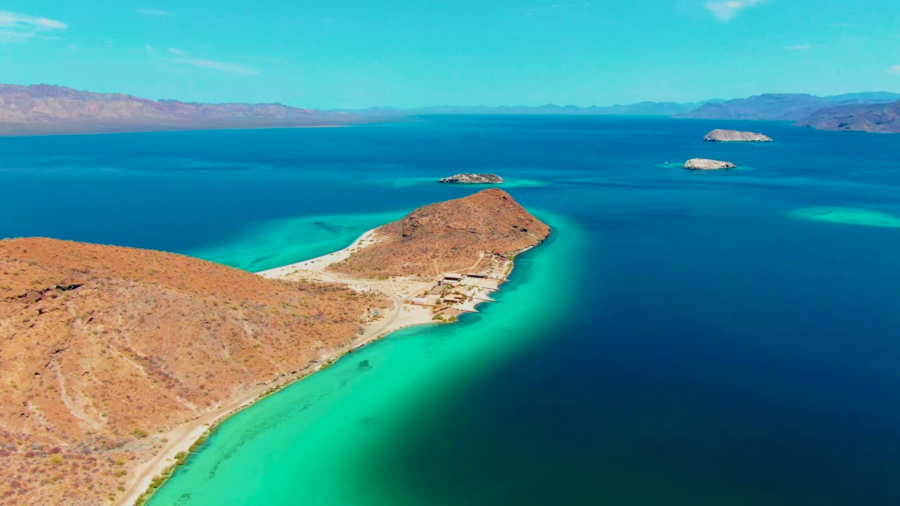 Opening aerial frame over Playa Santispac showing a rocky desert peninsula bordered by turquoise shallows, deep blue water beyond, distant islands, hazy mountains, and no visible people.