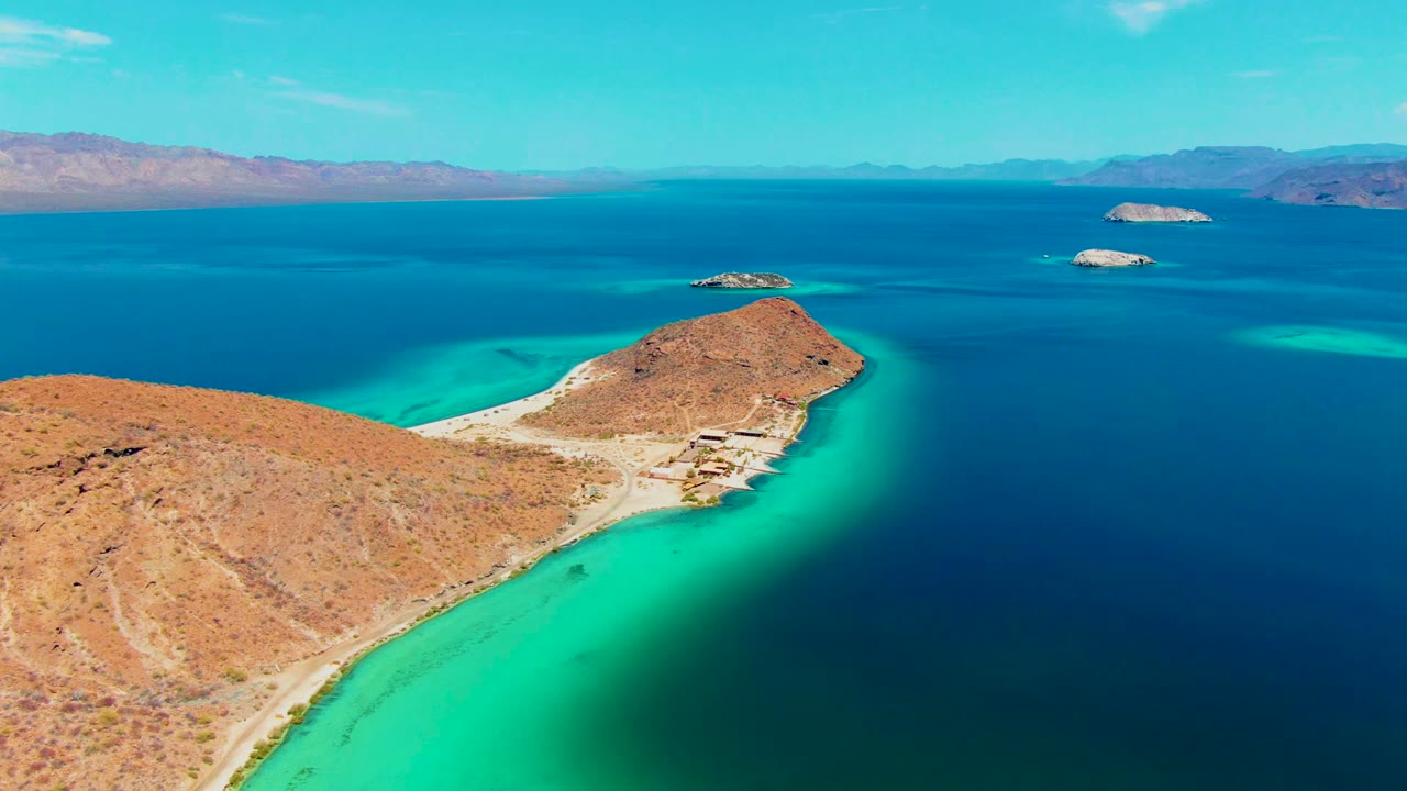 Desert coastline curves around turquoise water with small islands and mountains in the distance.