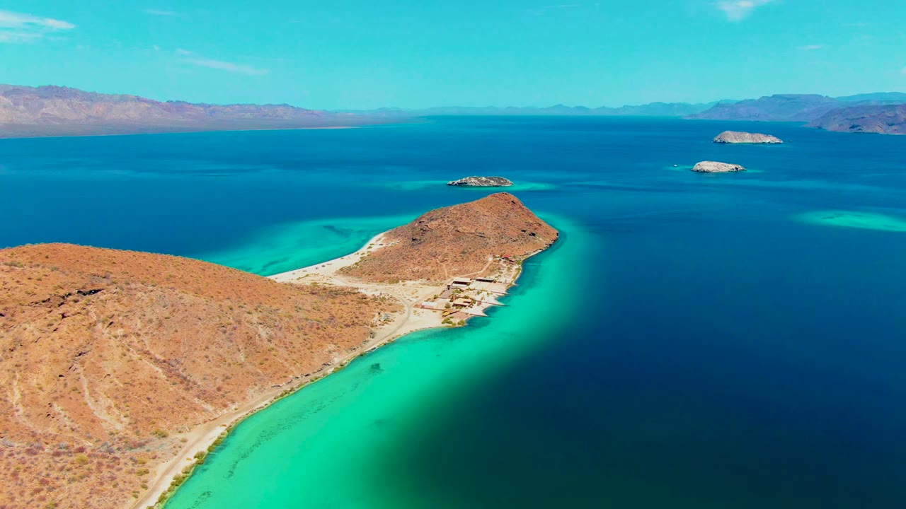 Aerial view over Playa Santispac with rocky ochre land in the lower frame, a narrow bright shoreline, turquoise water fading into deep blue, several distant islands, and no visible people.