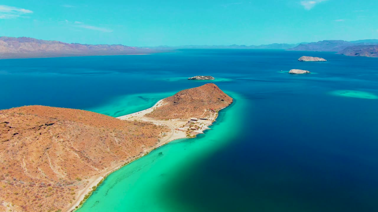 Aerial view over Playa Santispac showing an ochre desert peninsula, bright turquoise shallows, dark blue water, offshore islands, distant mountains, and no visible people.