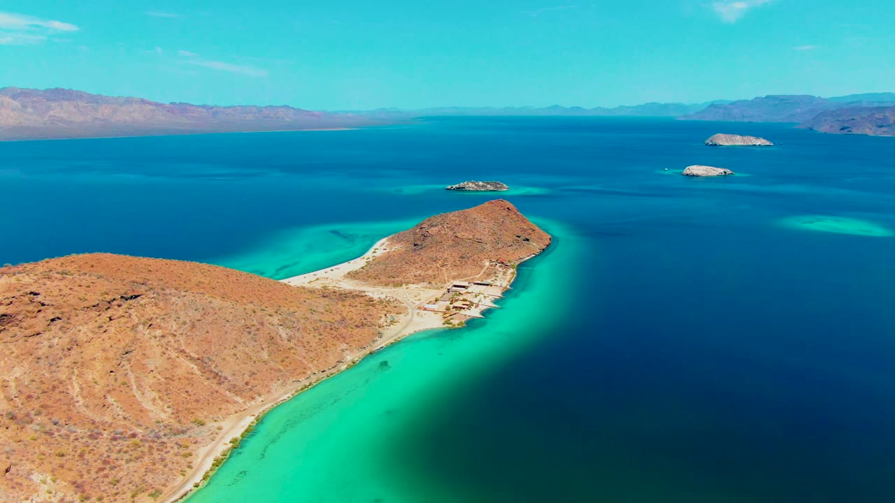 Aerial view of Playa Santispac with a rocky desert point in the lower center, luminous turquoise shallows along both sides, darker water beyond, islands, mountains, and no visible people.