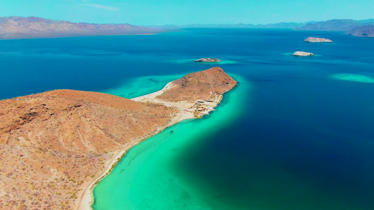 Aerial view at Playa Santispac, Baja California Sur, with a rocky peninsula in the foreground, bright turquoise shallows, dark blue open water, distant islands, and no visible people.