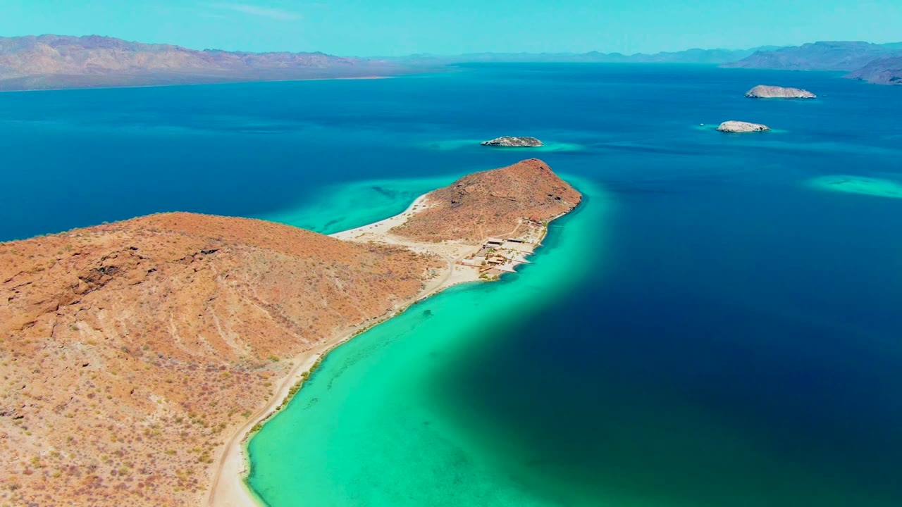 Aerial view over Playa Santispac with desert-colored rocky land at lower left, a pale beach line, turquoise water wrapping the shore, distant islands, and no visible people.